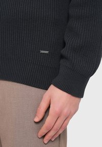 Σκούρο γκρι ριγέ πλεκτό πουλόβερ με εφαρμοστή γραμμή και μικρό α σημί λογότυπο στο κάτω αριστερό μέρος. Χέρι ακουμπισμένο σε ανοιχτό καφέ παντελόνι.