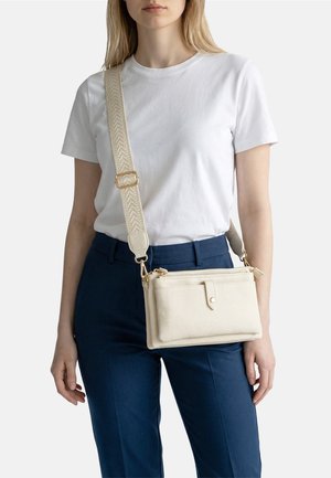 Donna che indossa una maglietta bianca e pantaloni blu scuro, con una borsa a tracolla in pelle beige con tracolla larga regolabile e tasca frontale.