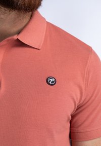 Polo shirt en corail clair fabriqué à partir d'un matériau texturé, doté d'un col standard et d'un badge logo noir sur la poitrine gauche.