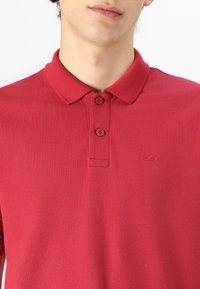 Sarkans polo krekls, izgatavots no strukturēta auduma, ar divu pogu apkakli un mazu izšūtu logo uz krūšu. Īsās piedurknes.