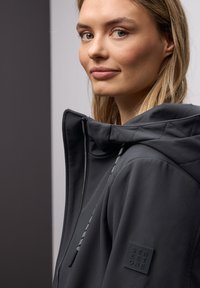 Jeune femme portant une veste à capuche foncée avec une fermeture éclair et des cordons, regardant légèrement sur le côté avec un fond neutre.