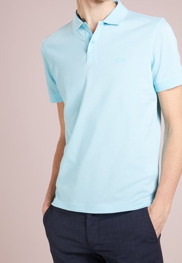 Polo shirt bleu clair en coton, avec une patte de boutonnage à trois boutons et un logo brodé discret sur la poitrine, associé à un pantalon sombre.