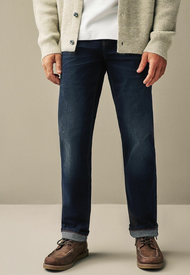 Donkerblauwe denim jeans met een rechte pasvorm, gecombineerd met een lichtgrijze geribde cardigan. Bruine veterschoenen, zichtbare plooien op de jeans.