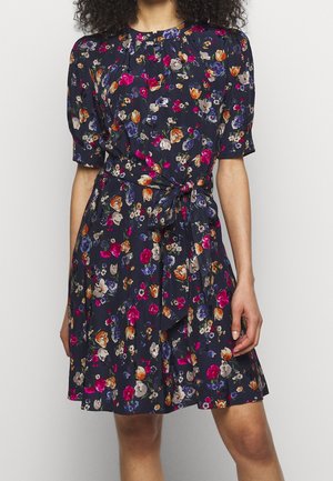 Vrouw draagt een marineblauwe bloemenjurk met korte pofmouwen, een taillekoord en kleurrijke bloemen in roze, oranje en paarse tinten.