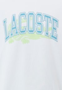 Balts T-krekls, kurā ar gaiši ziliem kontūrīgiem burtiem ir uzraksts "LACOSTE", zem kura atrodas zaļš aligatora attēls. Izgatavots no kokvilnas, ar minimālu tekstūru.