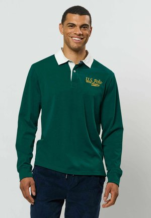 Homme souriant portant un polo à manches longues vert foncé avec col blanc et logo jaune U.S. Polo Assn. sur la poitrine.