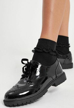Chaussures oxford en cuir verni noir avec des détails perforés et des lacets, assorties à des chaussettes noires côtelées avec des bords froncés.