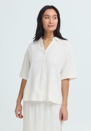 Kobieta o czarnych włosach, ubrana w luźną białą koszulę na guziki z krótkimi rękawami oraz pasującą białą spódnicę, patrząca lekko w bok.