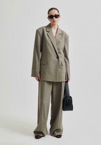 Överdimensionerad khaki-blazer och vida byxor i slät väv. Modellen håller en svart väska och har på sig svarta solglasögon. Enkel design, inga mönster.