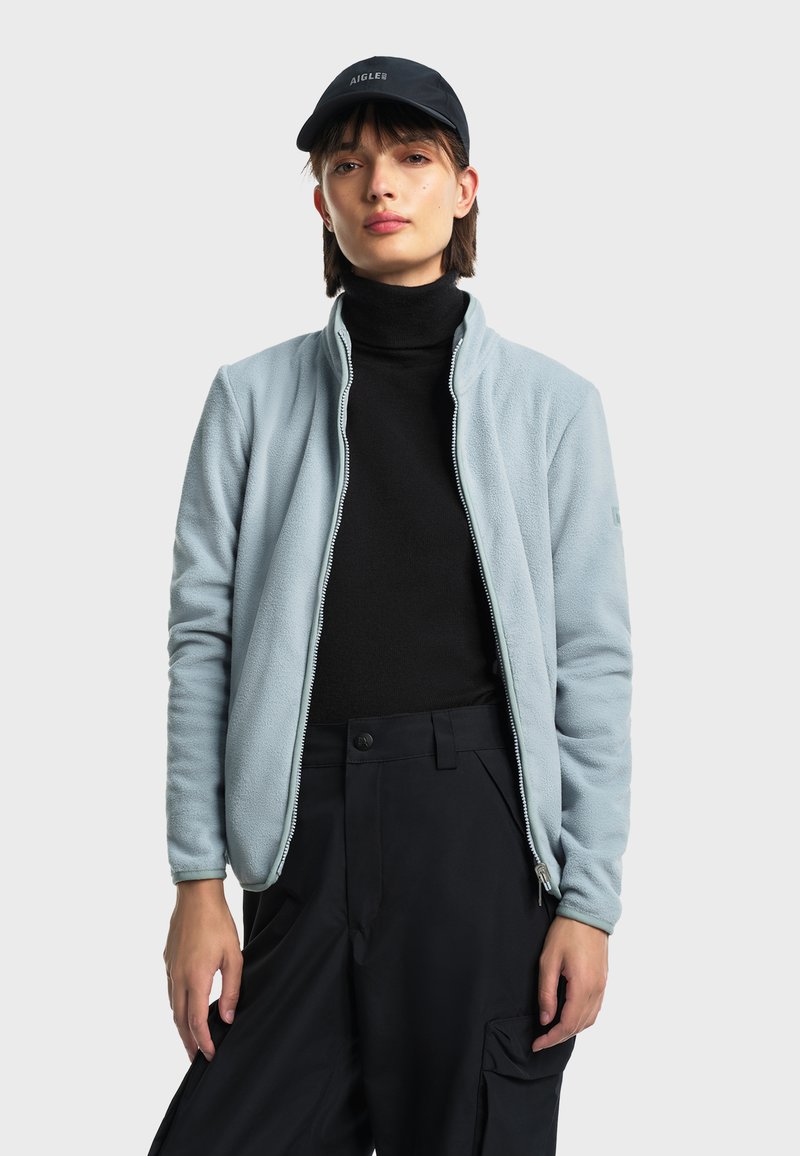 Svijetloplava flis jakna s zatvaranjem na patentni zatvarač i stojećim ovratnikom, nosi se preko crne majice s visokim ovratnikom. Uparen s tamnim cargo hlačama. Prikazana crna kapa.