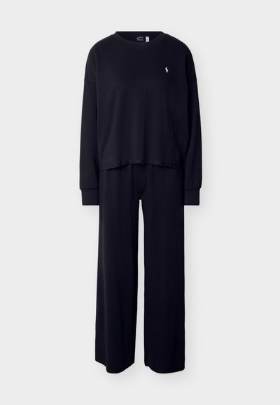 Camiseta negra de manga larga con un pequeño logo blanco en el pecho, combinada con pantalones negros de pierna ancha a juego sobre un fondo liso.