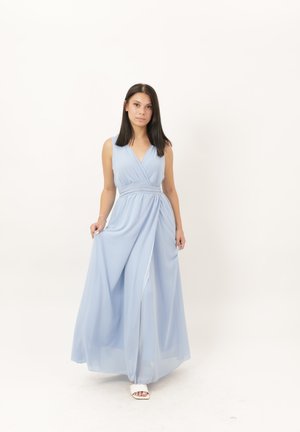 Frau mit langen dunklen Haaren, die ein ärmelloses hellblaues bodenlanges Kleid trägt, hält den Rock und steht vor einem schlichten weißen Hintergrund.