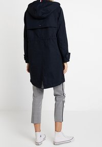 Impermeabile lungo blu navy con cappuccio e chiusura con bottoni, dotato di vita elasticizzata e spacco sul retro. Indossato con pantaloni corti a quadri e scarpe da ginnastica.