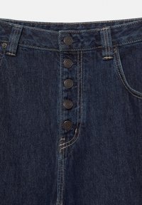Mørkeblå denimjeans med fem synlige sorte knapper på gylpen og kontrasterende hvide syninger omkring taljebåndet og lommerne.