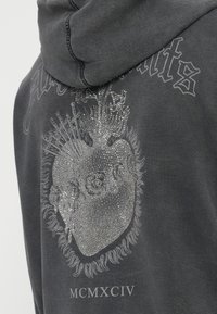 Σκούρα γκρι φούτερ με κουκούλα με το λαμπερό σχέ διο ενός ανατομικού καρδιάς διατρυπημένης από σπαθιά και φλόγες, με το λατινικό αριθμό MCMXCIV από κάτω.