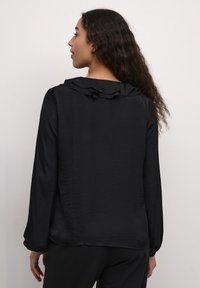 Black long-sleeve blouse with a ruffled collar and textured fabric, featuring a relaxed fit and slightly curved hem at the back.
