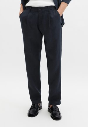 Mann trägt dunkelblaue, plissierte Hose mit schwarzen Lederloafern und Händen in den Taschen vor einfachem Hintergrund.