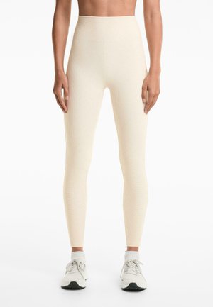 Világos bézs színű, magas derekú leggings, texturált, bordázott mintával. Fehér sportcipővel párosítva, fűzős elöl és fekete talppal.