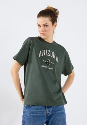 Oliven grøn bomulds t-shirt med korte ærmer, der har "ARIZONA" print og vestligt inspirerede grafikker, parret med blå denim jeans.
