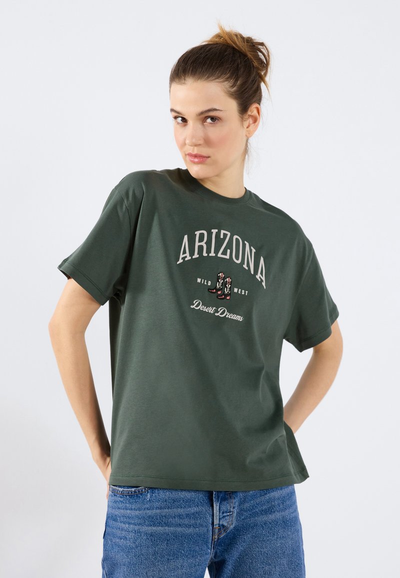 Oliivinvihreä puuvillapaita lyhyillä hihoilla, jossa on "ARIZONA" -painatus ja lännen teemaisia grafiikoita, yhdistettynä sinisiin farkkullareihin.