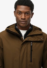 Brązowa wodoodporna kurtka z regulowanym kapturem, z przodu na zamek, czarnymi akcentami i logo na piersi; gładka tekstura i dopasowany krój.