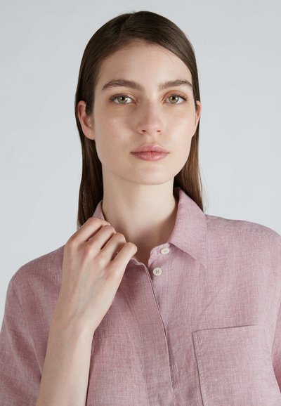 Camicia di lino rosa chiaro con maniche corte, colletto a punta e taschino sul petto. Tessuto strutturato con una vestibilità leggermente rilassata.