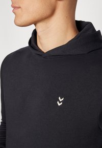 Mężczyzna w czarnej bluzie z kapturem z małym białym podwójnym logo chevron na piersi, pokazany od szyi do połowy torsu, zwrócony w lewo.