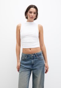 Weißes ärmelloses Crop-Top mit hohem Kragen, kombiniert mit hellblauen, locker sitzenden Jeans aus Denim mit subtilen Verwaschungen und fünf Taschen.