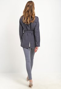 Szary płaszcz trencz z paskiem w talii, długimi rękawami i zakładką z tyłu. Noszony na szarych obcisłych dżinsach i jasnoszarych szpilkach.