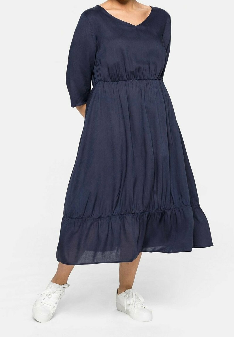 Femme portant une robe bleu marine mi-longue avec des manches trois-quarts et des baskets blanches, debout devant un fond uni.