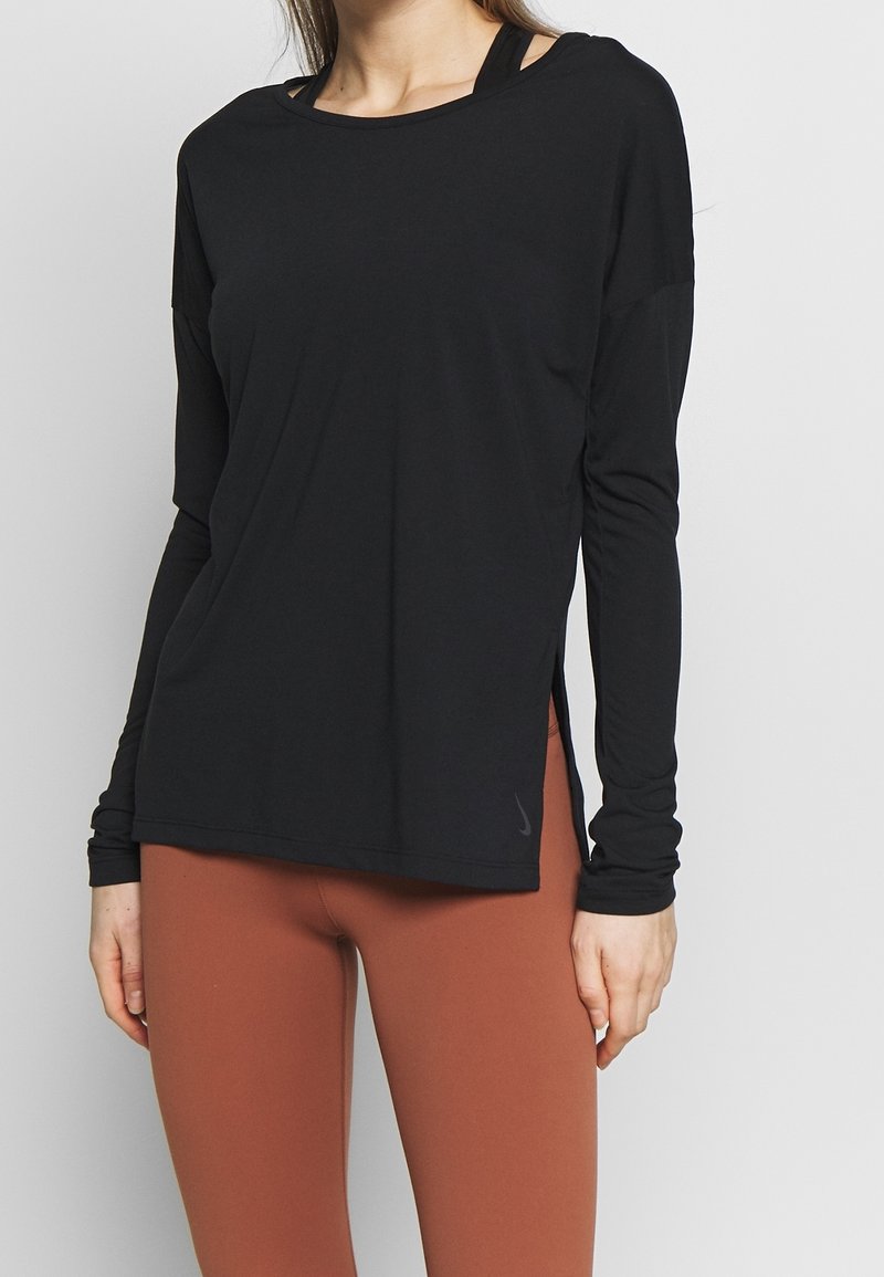 Top noir à manches longues en tissu léger, avec un col arrondi, des fentes latérales et un accent logo sur l'ourlet. Associé à un legging marron.