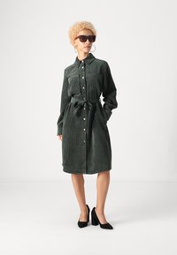 Σκοτεινό πράσινο πουκαμίσα-φόρεμα από χοντρό πανί με μακριά μανίκια, κουμπιά μπροστά, δύο τσέπες στο στήθος, ζώνη στη μέση και μήκος μέχρι το γόνατο. Μαύρες ψηλοτάκουνες γόβες.