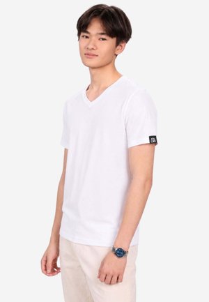 Giovane uomo in piedi, indossa una T-shirt bianca a V con toppa con logo sulla manica, pantaloni beige e un orologio d'argento con quadrante blu.
