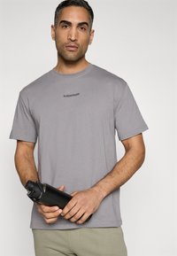 Tricou gri cu mânecă scurtă și decolteu rotund, cu un logo negru subtil. Modelul ține o sticlă de apă neagră elegantă.