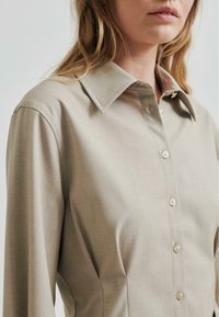Lys beige skjorte med struktured krage, lange ermer og subtil tekstur. Har en rad med perle-liknende knapper foran.