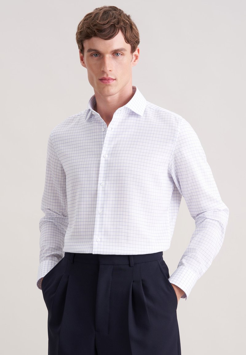 Chemise blanche à boutons avec un motif quadrillé bleu clair, tissu texturé, col pointu et manches longues. Portée avec un pantalon sombre ajusté.