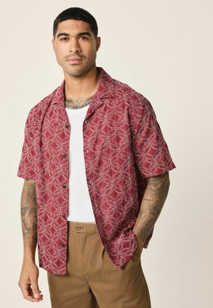 Short-sleeve red patterned shirt with a geometric design, light fabric, and black buttons, paired with a white tank top and beige pants.