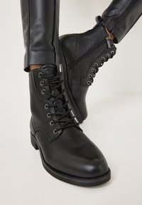 Botas de cuero negro hasta el tobillo con punta redonda, cordones en la parte delantera y cremallera lateral. Acabado texturizado y suela de goma resistente.