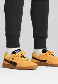 Geel suède sneakers met zwarte accenten, gestructureerde veters en een rubberen zool, gecombineerd met donkere trainingsbroeken en witte crew-sokken.