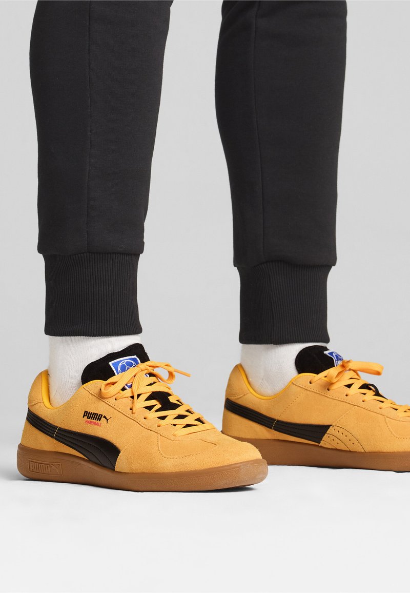 Geel suède sneakers met zwarte accenten, gestructureerde veters en een rubberen zool, gecombineerd met donkere trainingsbroeken en witte crew-sokken.