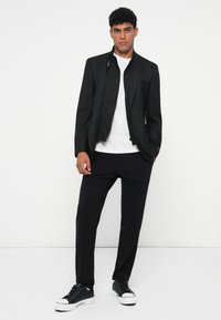 Svart skräddarsydd jacka med hög krage över en vit skjorta, i kombination med svarta byxor och svarta låga sneakers. Slätt tyg, minimalistisk design.