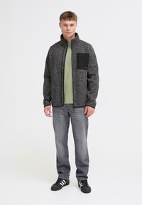 Herre grå fleecejakke med sort lynlås, høj krave og brystlomme. Båret over en grøn skjorte og parret med grå jeans og sorte sneakers.