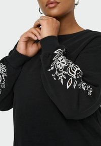 Czarna bluza z haftowanymi białymi kwiatowymi wzorami na rękawach. Materiał wydaje się miękki, z klasycznym okrągłym dekoltem i ściągaczami przy mankietach.