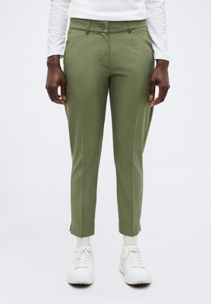 Persoon draagt olijfgroene, op maat gemaakte broek, wit shirt met lange mouwen, witte sokken en witte sneakers, staand tegen een effen achtergrond.