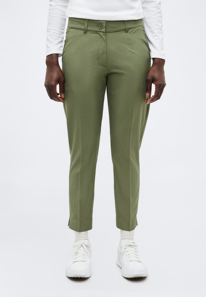 Άτομο που φοράει ελιά χρώματος εφαρμοστά παντελόνια, λευκό μακρυμάνικο τοπ, άσπρες κάλτσες και άσπρα αθλητικά παπούτσια, στέκεται μπροστά σε απλό φόντο.