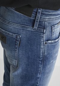 Plave traperice uskog kroja s teksturiranim završetkom, s crnom kožnom oznakom na pojasu i stražnjim zakrpa.