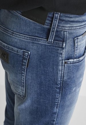 Vaqueros de mezclilla azul con un corte ajustado, que presentan un acabado texturizado, una etiqueta de cinturón de cuero negro y bolsillos traseros con parche.