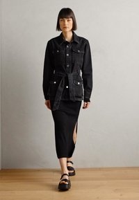 Svart denimjacka med framfickor och knappar, bärs över en svart åtsittande kjol med sidoklyfta, i kombination med kraftiga svarta sandaler.