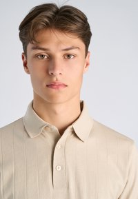 Junger Mann mit hellbraunen Haaren und haselnussbraunen Augen, der ein beiges, geripptes Poloshirt trägt und mit dem Gesicht nach vorne vor einem schlichten, hellen Hintergrund steht.