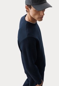 Sudadera texturizada en color navy con un patrón de espiga, cuello acanalado y puños ajustados, combinada con una gorra gris.